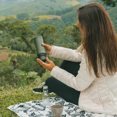 atmosphärisches Bild mit viel grüner Landschaft im Hintergrund und im Vordergrund auf einer Decke sitzend eine Frau, die mit der portable Espressomaschine OutIn Nano einen Espresso zubereitet